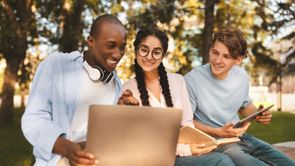 Excited international students studying in park, enjoying the results of their job, looking at laptop screen and smiling, sitting together on bench in campus.